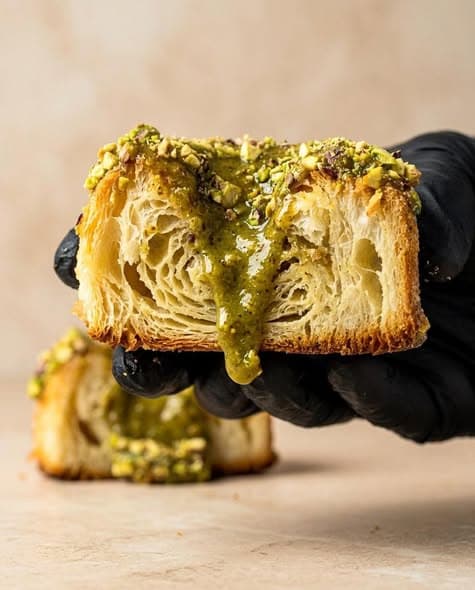 A pistachio pastry cut open in a gloved hand, pistachio cream running down its honeycomb layers